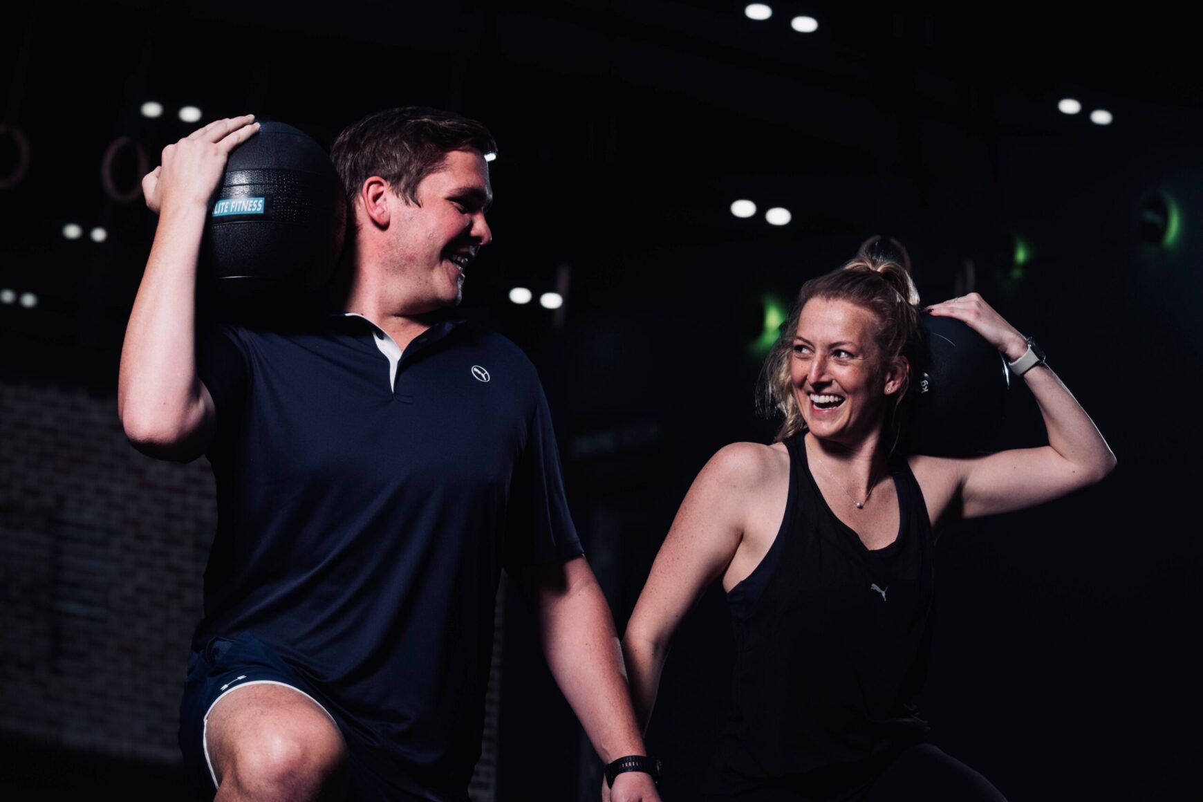 Two people enjoying a group training session in a gym, holding medicine balls and smiling